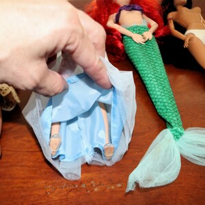 May include: A doll wearing a light blue skirt with a sheer tulle overlay, positioned next to a mermaid doll with red hair and a green scale-patterned tail, and a doll with dark hair wearing white underwear, all featuring articulated limbs.
