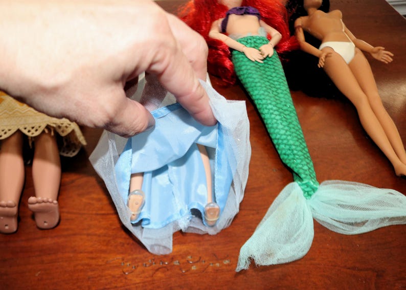 May include: A doll wearing a light blue skirt with a sheer tulle overlay, positioned next to a mermaid doll with red hair and a green scale-patterned tail, and a doll with dark hair wearing white underwear, all featuring articulated limbs.