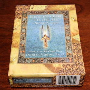 May include: A rectangular box set titled Healing with the Angels Oracle Cards by Doreen Virtue, Ph.D. The box features a central blue panel with an angel illustration, surrounded by a decorative border and golden-toned artwork. Includes a 44-card deck.