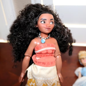 May include: A doll with long, voluminous dark brown curly hair, brown eyes, and a tan complexion. The doll wears an orange patterned top with a blue stone pendant necklace and a cream and yellow skirt with a geometric star pattern on the bottom.