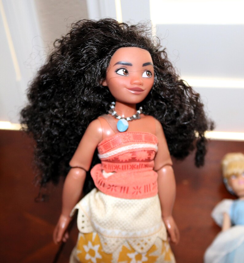 May include: A doll with long, voluminous dark brown curly hair, brown eyes, and a tan complexion. The doll wears an orange patterned top with a blue stone pendant necklace and a cream and yellow skirt with a geometric star pattern on the bottom.