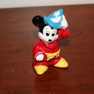 May include: A ceramic figurine of Mickey Mouse dressed as a sorcerer. He is wearing a red robe with yellow accents, a blue hat with a white star, and black shoes.