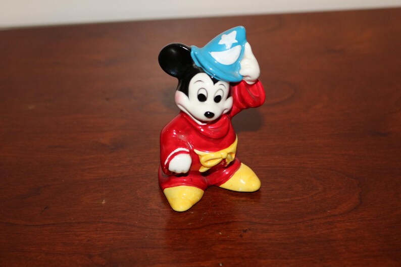 May include: A ceramic figurine of Mickey Mouse dressed as a sorcerer. He is wearing a red robe with yellow accents, a blue hat with a white star, and black shoes.