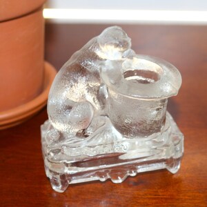 May include: A clear glass figurine of a cat sitting on a base with a hat. The cat is looking down at the hat, which has a small hole in the top.