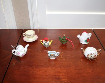 May include: A collection of seven miniature ceramic items including a teacup and saucer, a white teapot with pink flowers, a yellow and black teapot, a green Christmas tree ornament, a white pitcher, a white watering can, and a white teapot.