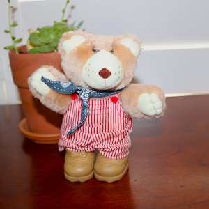 May include: A plush brown teddy bear wearing red and white striped overalls and a blue bandana. The bear is standing on a wooden surface.