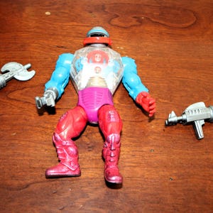 May include: A vintage action figure featuring a clear plastic chest plate with visible internal gears, blue arms, a purple waist, and red legs. The figure includes a silver axe accessory and a silver blaster accessory, with a red visor over the head.