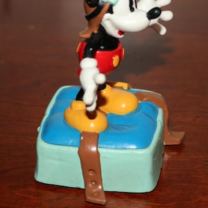 May include: A Mickey Mouse figurine in a pilot outfit, standing on a blue and green box. The figure has yellow shoes, red shorts, a black shirt, and a brown pilot hat. The box has a brown strap.