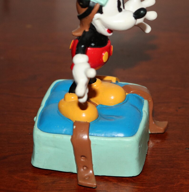 May include: A Mickey Mouse figurine in a pilot outfit, standing on a blue and green box. The figure has yellow shoes, red shorts, a black shirt, and a brown pilot hat. The box has a brown strap.