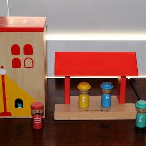 May include: A wooden toy set featuring a tall rectangular building with a red helipad roof, a red-roofed station platform, and four small wooden peg figures in red, yellow, blue, and teal, each wearing a painted tie and a matching hat.