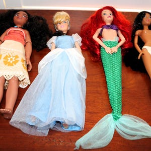 May include: Four dolls arranged in a row. The first has long dark curly hair, a red top, and a white patterned skirt. The second has blonde hair and a light blue gown. The third has long red hair and a green mermaid tail. The fourth has long black hair and white underwear.