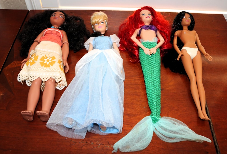 May include: Four dolls arranged in a row. The first has long dark curly hair, a red top, and a white patterned skirt. The second has blonde hair and a light blue gown. The third has long red hair and a green mermaid tail. The fourth has long black hair and white underwear.