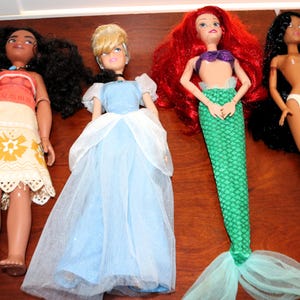 May include: Four dolls arranged in a row. The first has long dark curly hair, a red top, and a patterned cream skirt. The second has blonde hair, a blue dress with white sheer sleeves. The third has red hair and a green mermaid tail. The fourth has long black hair and white underwear.