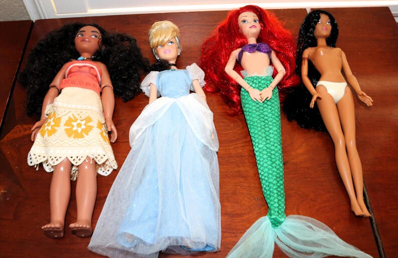 May include: Four dolls arranged in a row. The first has long dark curly hair, a red top, and a patterned cream skirt. The second has blonde hair, a blue dress with white sheer sleeves. The third has red hair and a green mermaid tail. The fourth has long black hair and white underwear.
