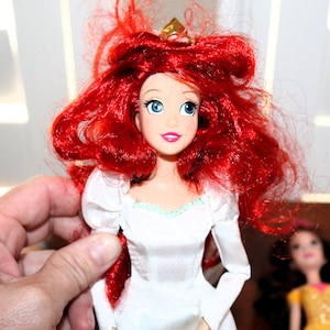 May include: A doll with bright red hair and blue eyes, wearing a white dress with a gold crown. The doll has a smiling expression and is held by a person's hand. Another doll is visible in the background.