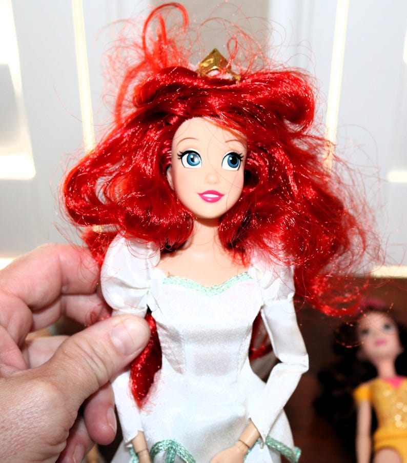 May include: A doll with bright red hair and blue eyes, wearing a white dress with a gold crown. The doll has a smiling expression and is held by a person's hand. Another doll is visible in the background.