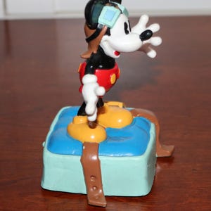 May include: A Mickey Mouse figurine in a pilot outfit, featuring a black body, white face, and red shorts. The figure stands on a blue base with brown straps and yellow shoes. The figurine is wearing a brown aviator helmet and goggles.
