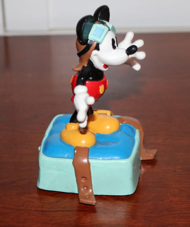 May include: A Mickey Mouse figurine in a pilot outfit, featuring a black body, white face, and red shorts. The figure stands on a blue base with brown straps and yellow shoes. The figurine is wearing a brown aviator helmet and goggles.