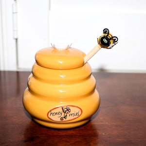 May include: A yellow ceramic honey pot with a lid and a wooden honey dipper. The pot is shaped like a beehive and has the words "honey miel" on the front. The dipper has a bee-shaped handle.