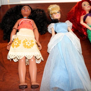 May include: Three dolls featuring a character with long dark curly hair in a tan and orange patterned outfit, a character with blonde hair in a light blue gown with a sheer overlay, and a character with long red hair and a green mermaid tail.