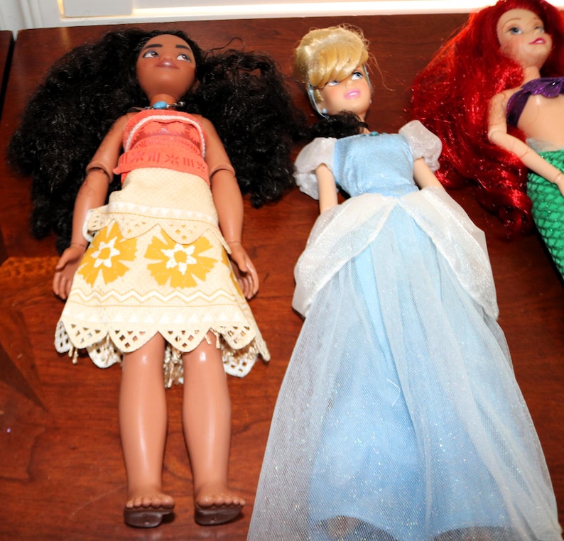 May include: Three dolls featuring a character with long dark curly hair in a tan and orange patterned outfit, a character with blonde hair in a light blue gown with a sheer overlay, and a character with long red hair and a green mermaid tail.