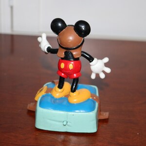 May include: A Mickey Mouse figurine in a pilot outfit, standing on a blue and green base. The figure has a brown helmet, red shorts, yellow shoes, and white gloves. The figurine is a collectible item.