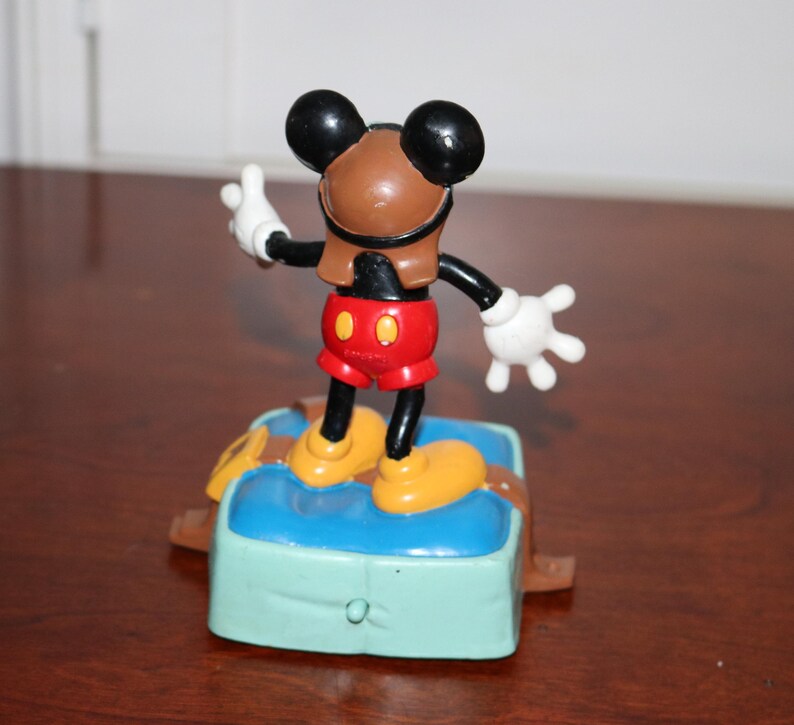 May include: A Mickey Mouse figurine in a pilot outfit, standing on a blue and green base. The figure has a brown helmet, red shorts, yellow shoes, and white gloves. The figurine is a collectible item.