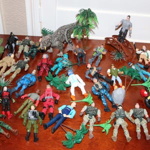 May include: A collection of toy action figures and plastic dinosaurs are arranged on a wooden surface. The figures are in various poses and outfits, with green palm tree decorations. The color palette includes red, blue, green, and tan.
