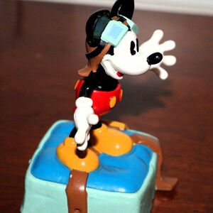 May include: A colorful Mickey Mouse figurine in a pilot outfit, with goggles and a red shirt. The figure is standing on a blue and green box with brown straps. The figurine is a collectible item.