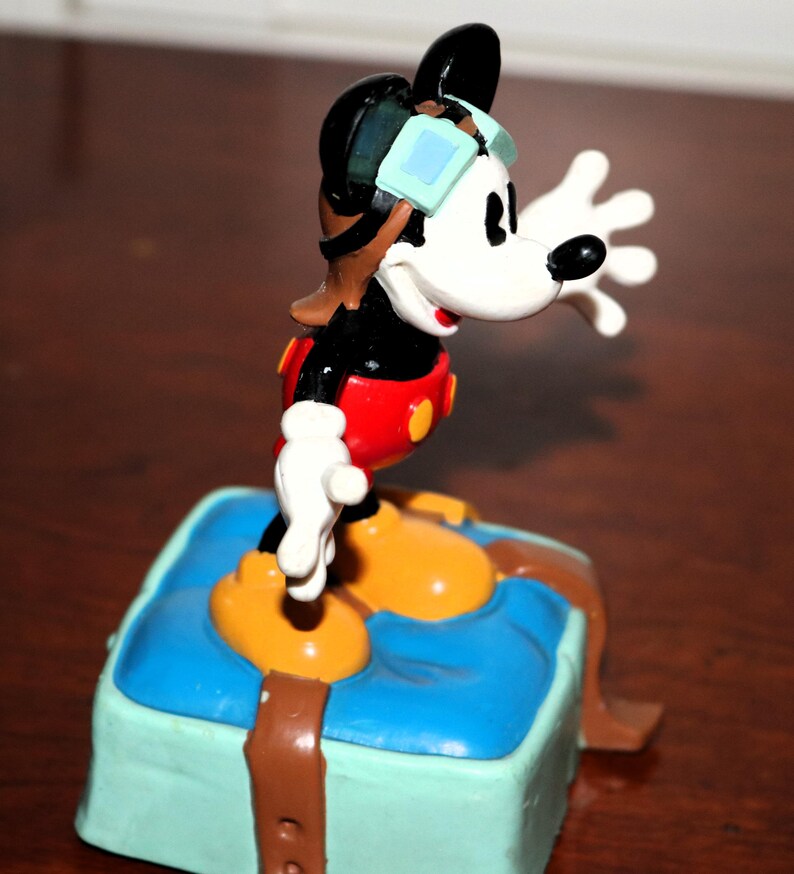 May include: A colorful Mickey Mouse figurine in a pilot outfit, with goggles and a red shirt. The figure is standing on a blue and green box with brown straps. The figurine is a collectible item.