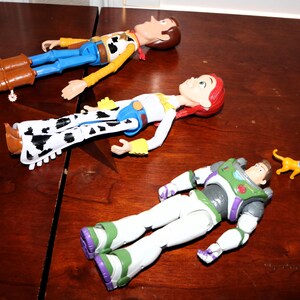 Puede incluir: Tres figuras de personajes de Toy Story: Woody con un atuendo de vaquero, Jessie con un atuendo de vaquera y Buzz Lightyear con un traje espacial, junto con una peque&ntilde;a figura de gato amarillo, todo sobre una superficie de madera.