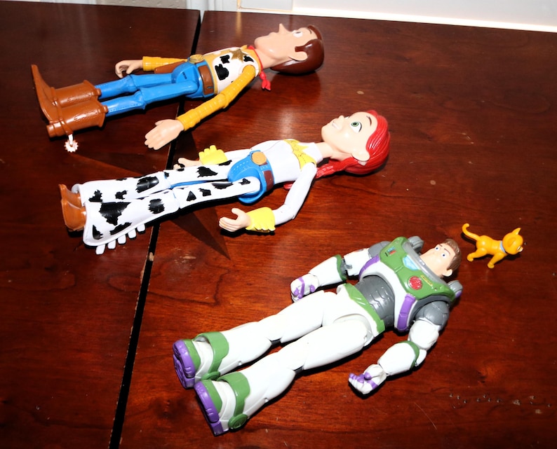 Puede incluir: Tres figuras de personajes de Toy Story: Woody con un atuendo de vaquero, Jessie con un atuendo de vaquera y Buzz Lightyear con un traje espacial, junto con una peque&ntilde;a figura de gato amarillo, todo sobre una superficie de madera.