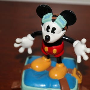May include: A Mickey Mouse figurine with black ears, a white face, and red shorts. The figure wears goggles and yellow shoes, standing on a blue and brown base. The figure's arms are outstretched.