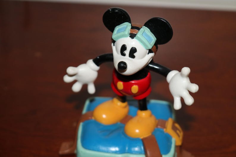 May include: A Mickey Mouse figurine with black ears, a white face, and red shorts. The figure wears goggles and yellow shoes, standing on a blue and brown base. The figure's arms are outstretched.
