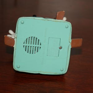 May include: A close-up of the bottom of a teal-colored toy with brown accents. The toy has a speaker grill and a battery compartment. The toy is on a dark brown wooden surface.