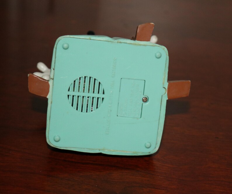 May include: A close-up of the bottom of a teal-colored toy with brown accents. The toy has a speaker grill and a battery compartment. The toy is on a dark brown wooden surface.