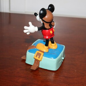 May include: A Mickey Mouse figurine in a pilot outfit stands on a light blue box with a brown strap and buckle. Mickey wears a brown helmet, red shorts, yellow shoes, and white gloves. The figurine is a collectible item.