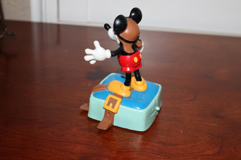 May include: A Mickey Mouse figurine in a pilot outfit stands on a light blue box with a brown strap and buckle. Mickey wears a brown helmet, red shorts, yellow shoes, and white gloves. The figurine is a collectible item.
