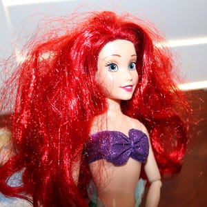 May include: A doll featuring long, vibrant red hair, light skin, and blue eyes. The doll wears a purple, glittery, strapless top with a bow detail at the center. The facial features include defined eyebrows, long eyelashes, and a soft pink smile.