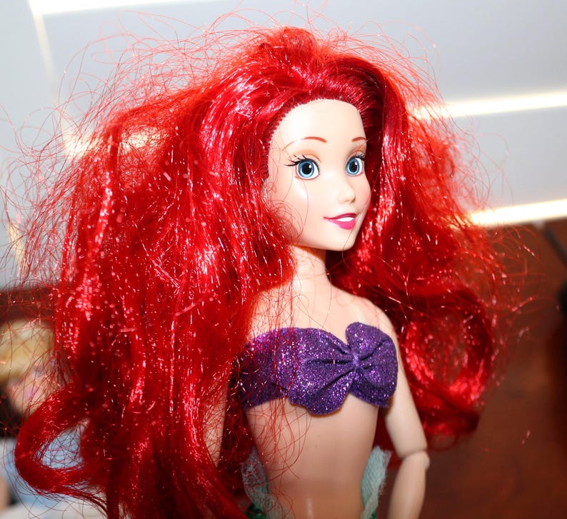 May include: A doll featuring long, vibrant red hair, light skin, and blue eyes. The doll wears a purple, glittery, strapless top with a bow detail at the center. The facial features include defined eyebrows, long eyelashes, and a soft pink smile.