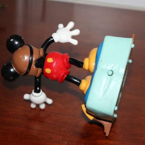 May include: A vintage Mickey Mouse alarm clock. The clock features Mickey's iconic brown head, black ears, red shorts, and white gloves. The clock's face is a light blue color with a brown base.