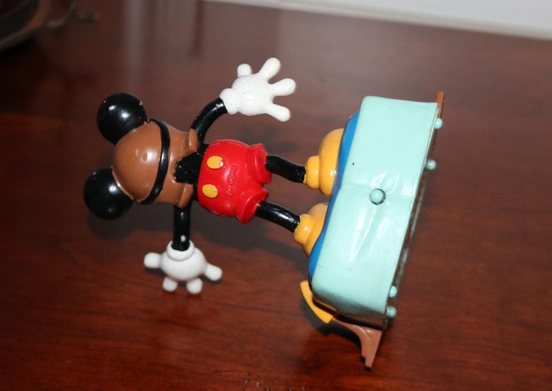May include: A vintage Mickey Mouse alarm clock. The clock features Mickey's iconic brown head, black ears, red shorts, and white gloves. The clock's face is a light blue color with a brown base.