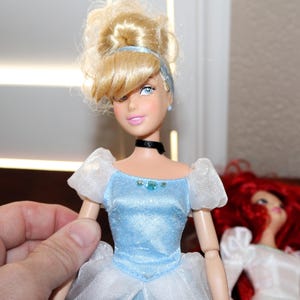 May include: A Cinderella doll with blonde hair styled in a bun, wearing a light blue dress with white accents. The doll has blue eyes, pink lips, and a black choker. Another doll with red hair is visible in the background.