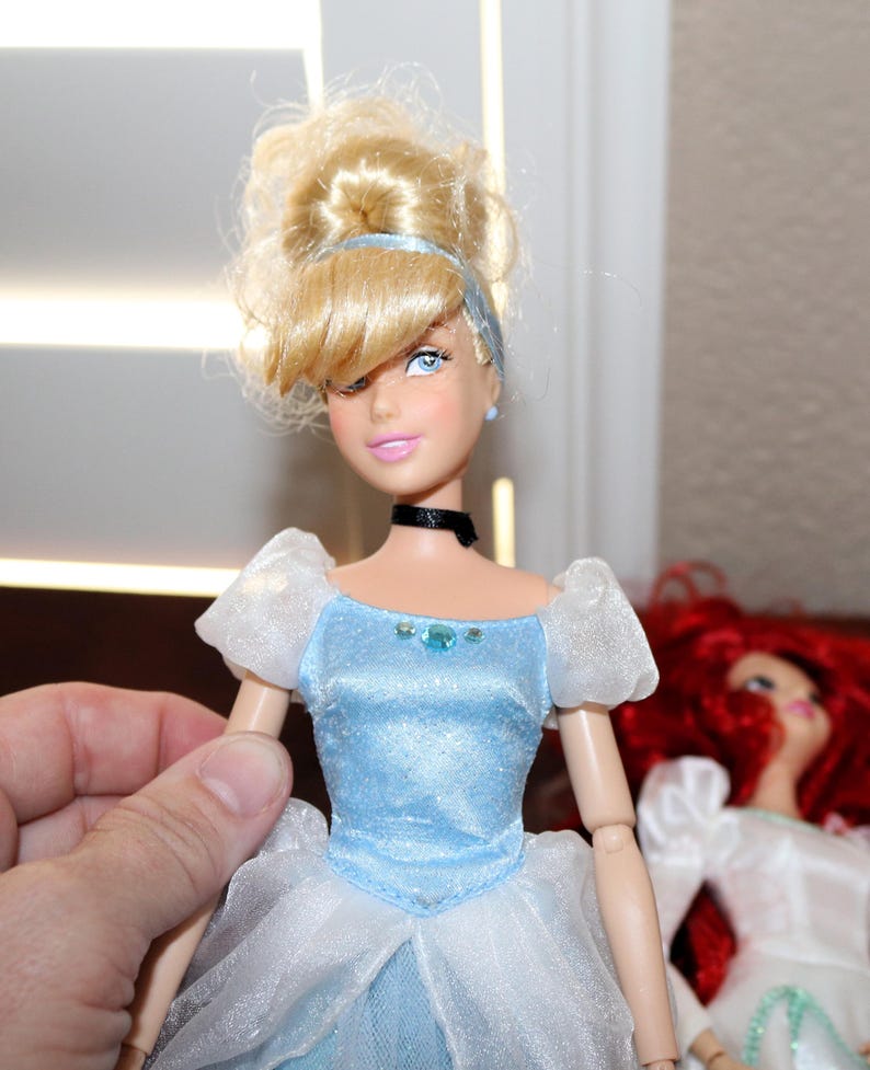 May include: A Cinderella doll with blonde hair styled in a bun, wearing a light blue dress with white accents. The doll has blue eyes, pink lips, and a black choker. Another doll with red hair is visible in the background.