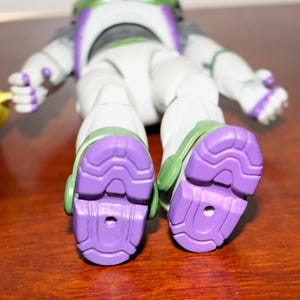 Puede incluir: Primer plano de los pies de una figura de acci&oacute;n de Buzz Lightyear. La figura es principalmente blanca con detalles verdes y morados. Las suelas de las botas son de un morado texturizado, y la figura est&aacute; sobre una superficie marr&oacute;n.