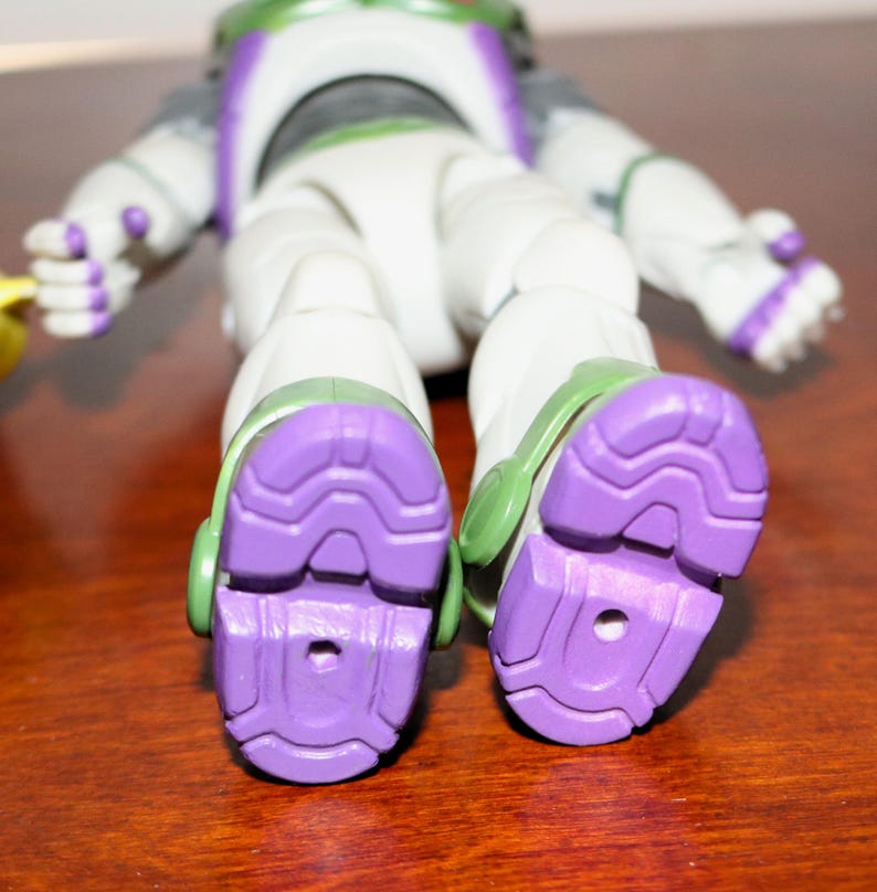 Puede incluir: Primer plano de los pies de una figura de acci&oacute;n de Buzz Lightyear. La figura es principalmente blanca con detalles verdes y morados. Las suelas de las botas son de un morado texturizado, y la figura est&aacute; sobre una superficie marr&oacute;n.