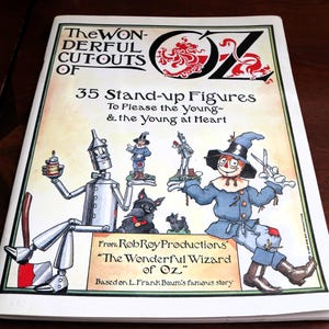May include: A book titled "The Wonderful Cutouts of Oz" featuring illustrations of characters from the Wizard of Oz. The cover art includes the Tin Man, Scarecrow, and Toto, with text that reads "35 Stand-up Figures".