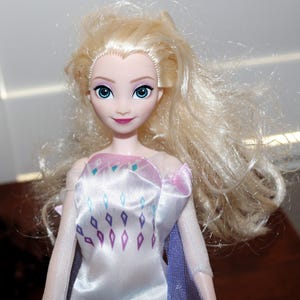 May include: A doll with long, wavy blonde hair and blue eyes. The doll is wearing a white dress with a pink and purple bodice and a purple cape. The dress has a diamond pattern in shades of blue and purple.