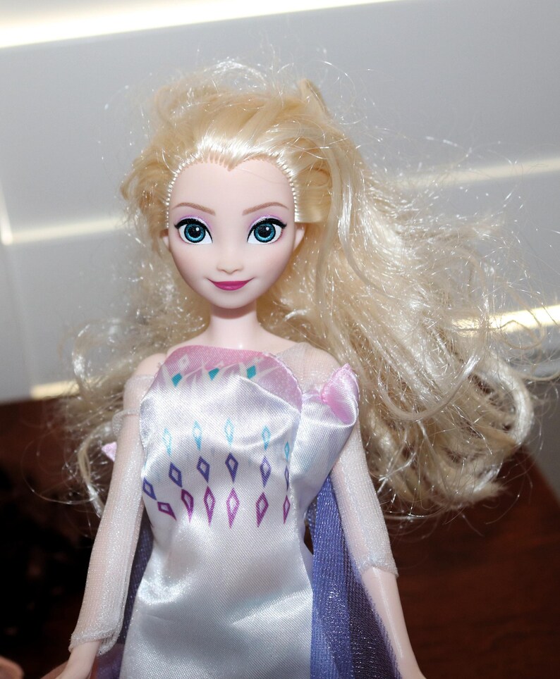 May include: A doll with long, wavy blonde hair and blue eyes. The doll is wearing a white dress with a pink and purple bodice and a purple cape. The dress has a diamond pattern in shades of blue and purple.