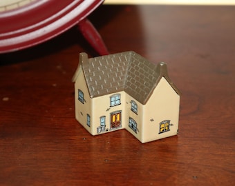 May include: A miniature ceramic house with a tan exterior and a brown roof. The house features detailed windows, a front door, and a chimney. The house is a decorative item.